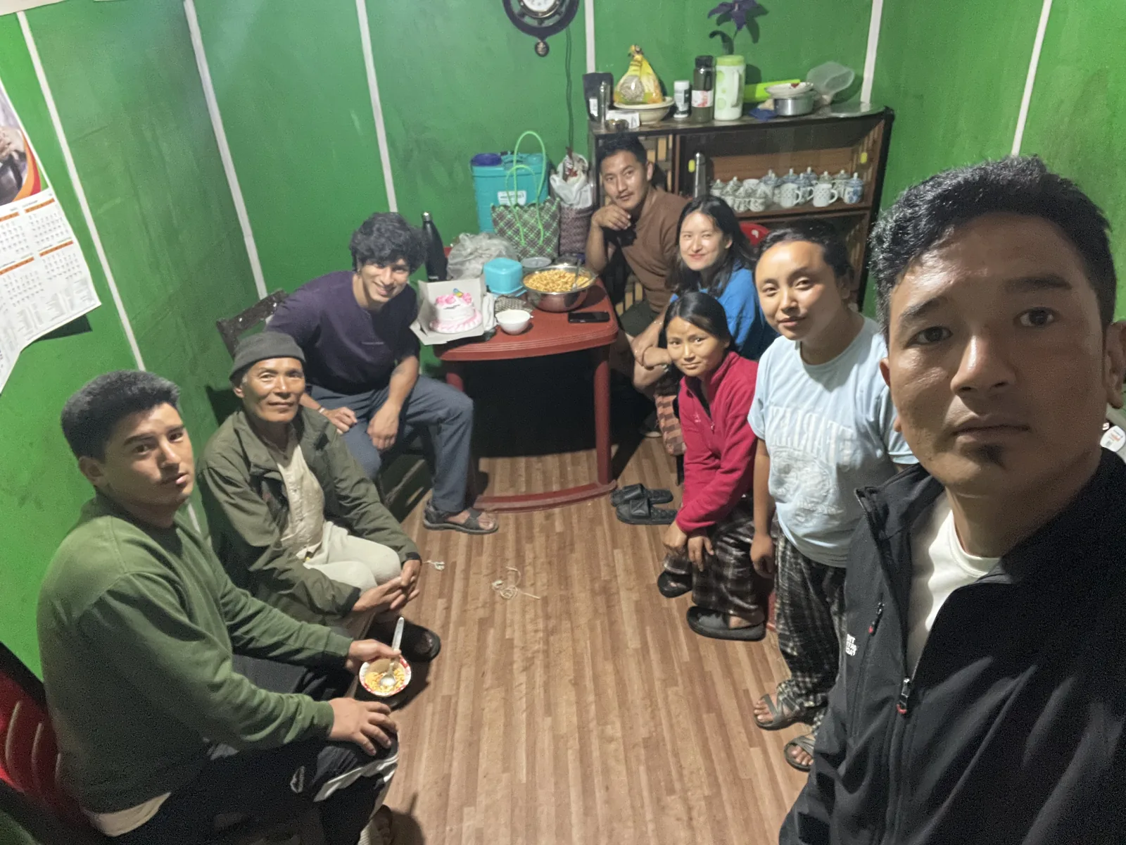 Hosts and guests crowded into a small green-walled room around a low table of food, one taking the photo