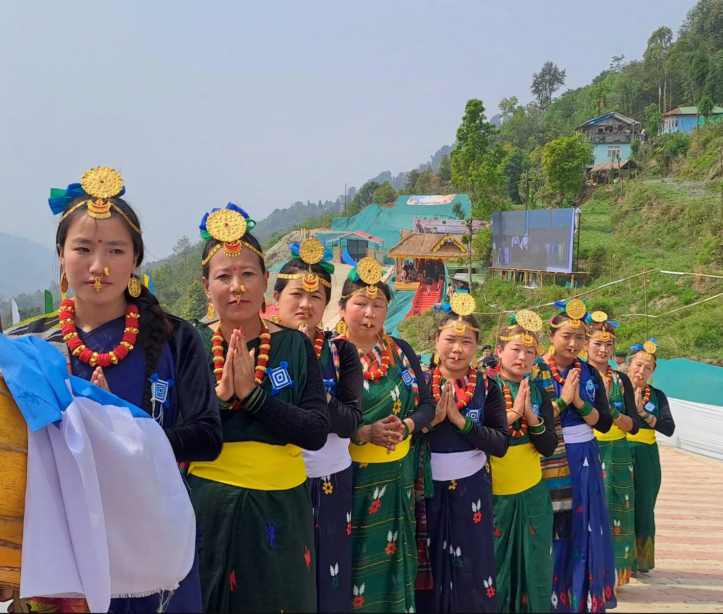 Women in traditional dress standing in a line with hands folded in welcome at the village entrance