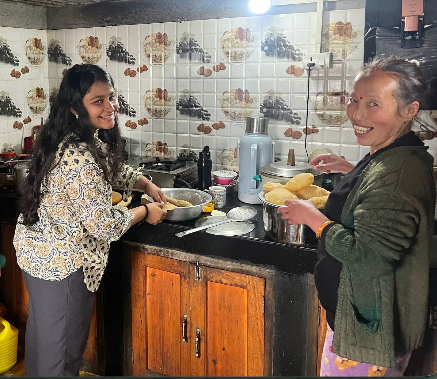 A guest rolling dough beside didi at the stove, both laughing, puris stacked on a plate
