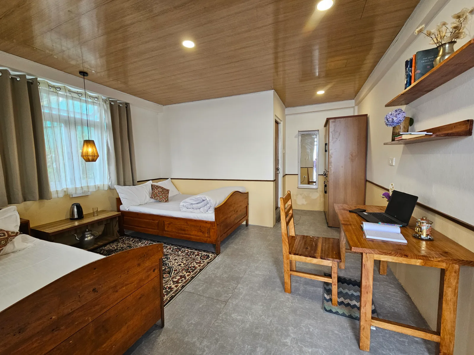 A room at Kothey Bari homestay: wooden ceiling, two beds, bookshelves and a desk with a laptop and kettle
