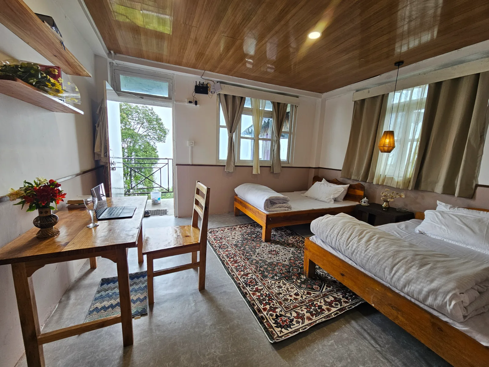 A room at Yakten homestay: two wooden beds, a long desk with a laptop and flowers, and a door open onto the balcony